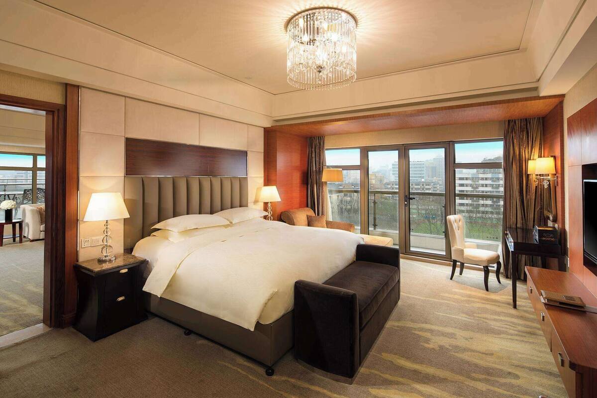 A spacious hotel room with a large bed, two bedside tables, a chandelier, a TV, and a view of the city.