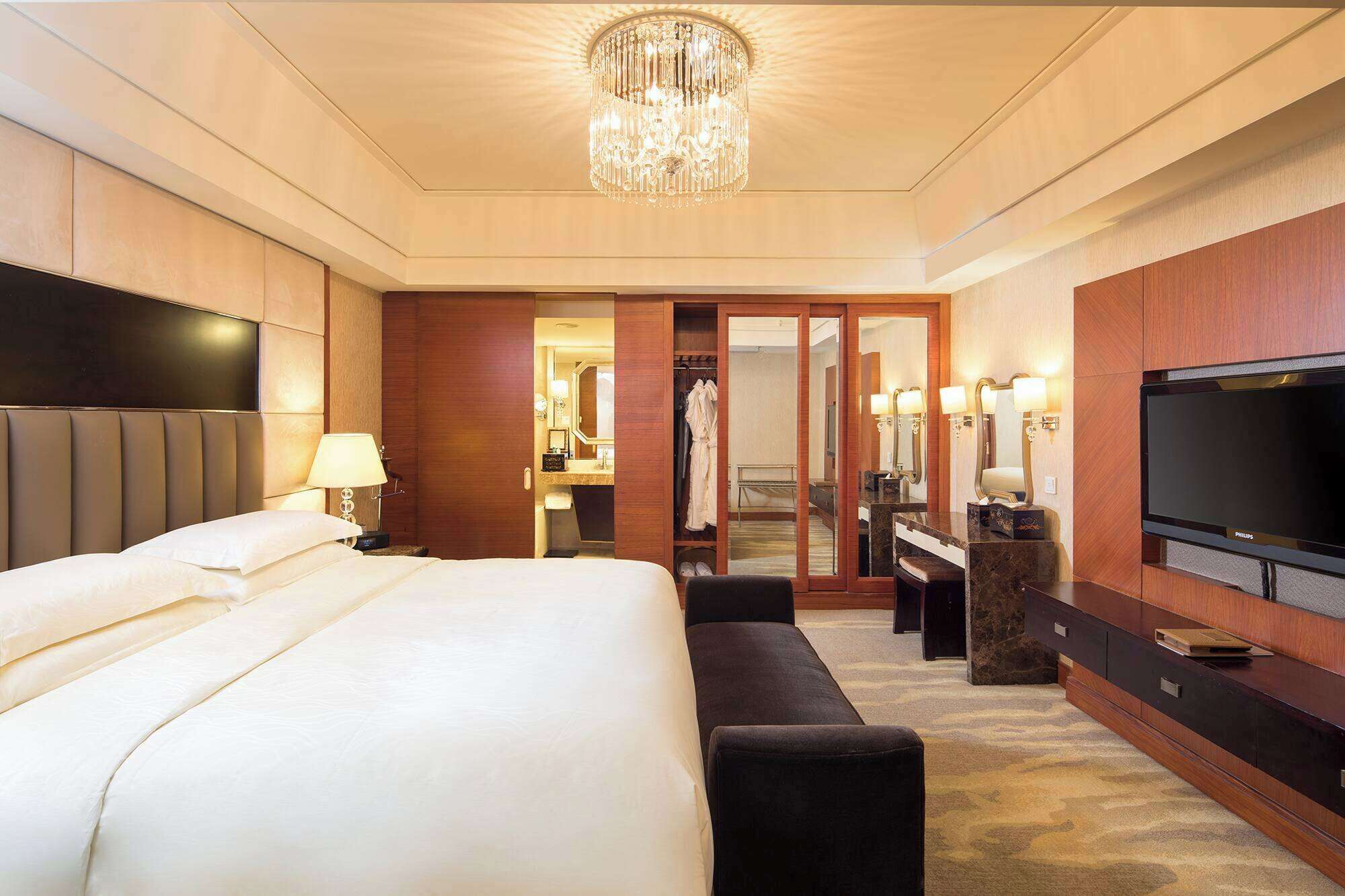 A hotel room with a large bed, a TV, a mirror, and a chandelier.