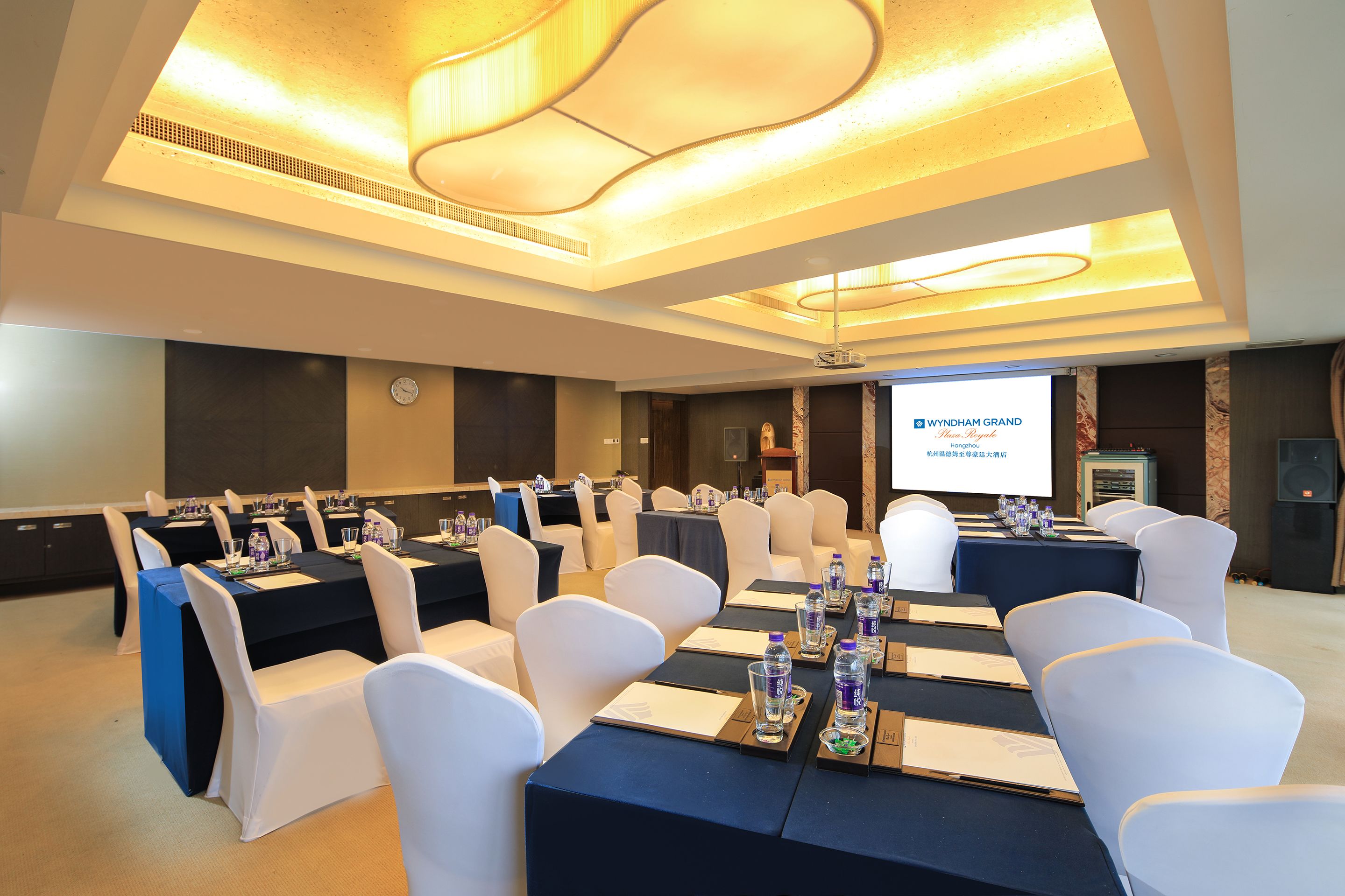 A conference room with multiple tables, white chairs, and a projector screen displaying 'Wyndham Grand'.