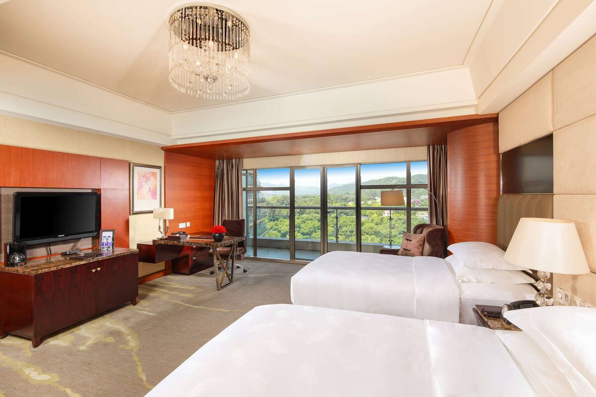 A hotel room with a large TV, a desk, two beds, and a view of the outdoors.