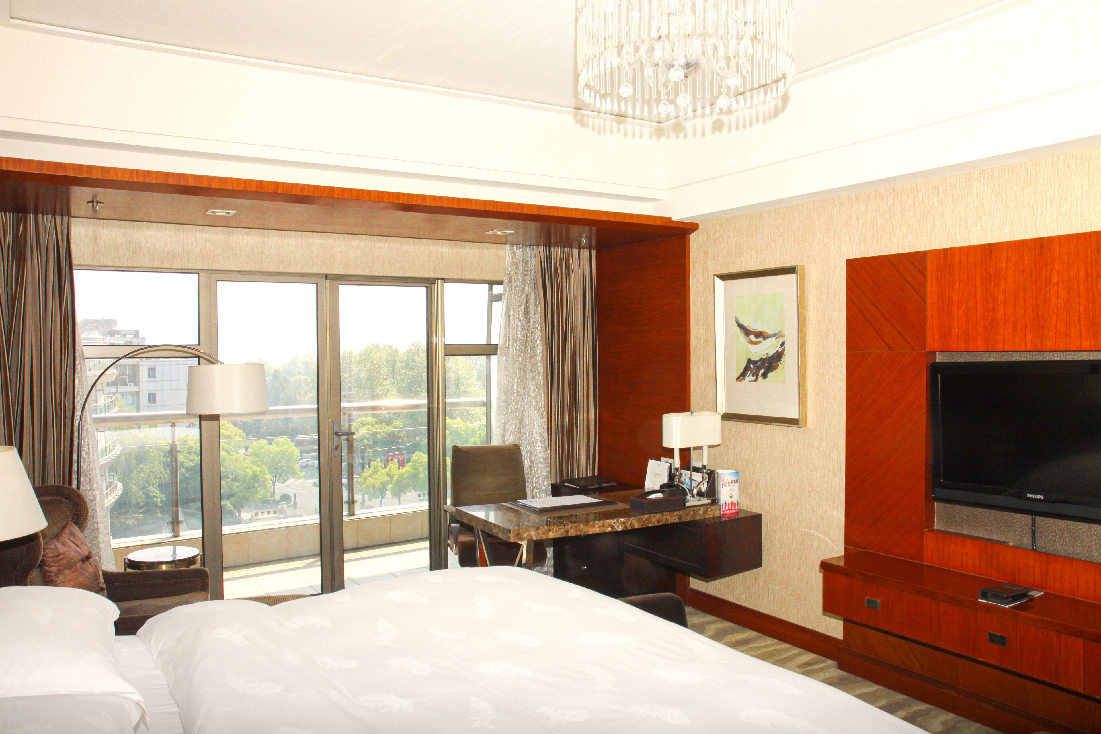 A hotel room with a large bed, a desk with a computer, a television, and a view of the city through large windows.