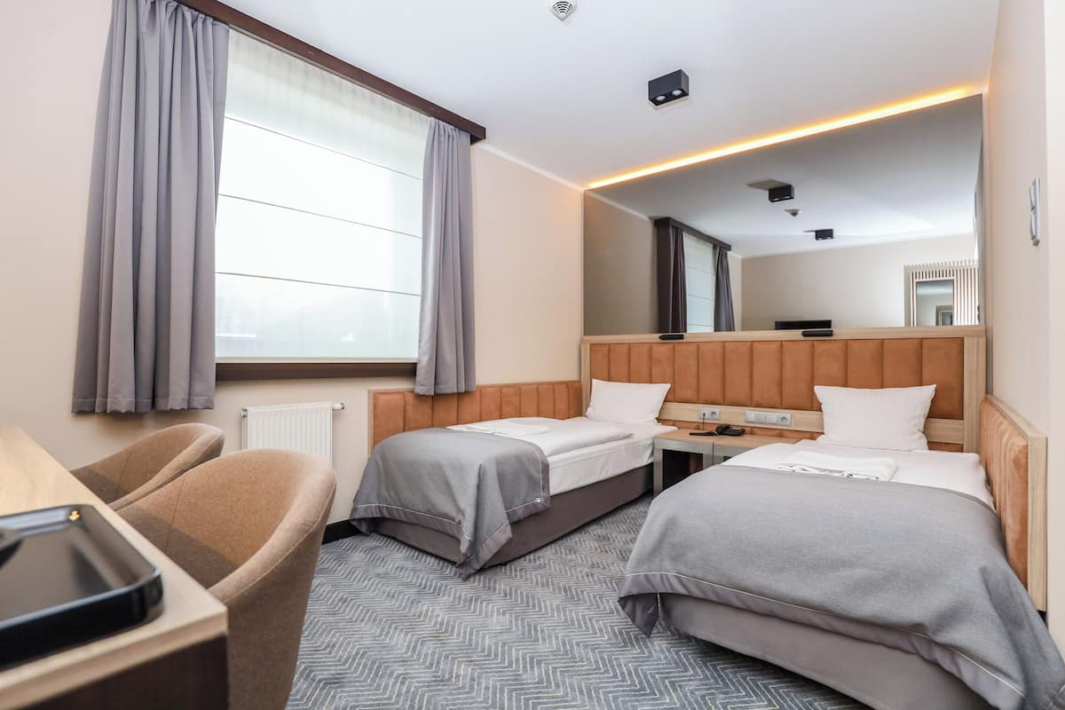 A hotel room with two beds, a desk with a chair, a large window with curtains, and a mirror above the bed.