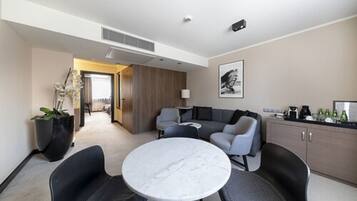 Una habitación de hotel moderna con una mesa de mármol, sofás grises, una zona de cocina y una planta grande en maceta.