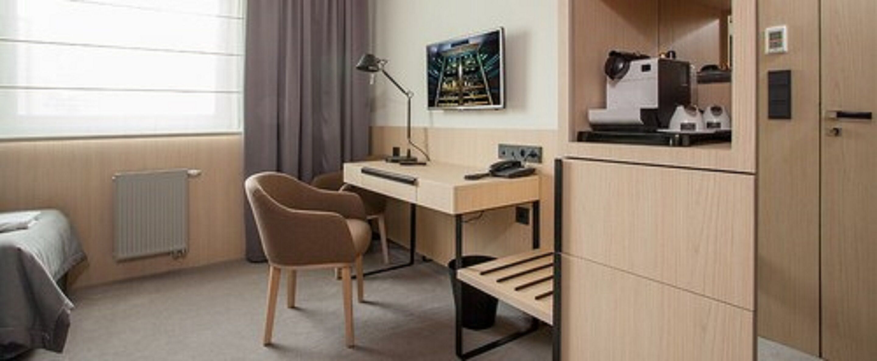 Quarto de hotel com escrivaninha, duas cadeiras, radiador, janela com cortinas, um quadro na parede e um minibar.