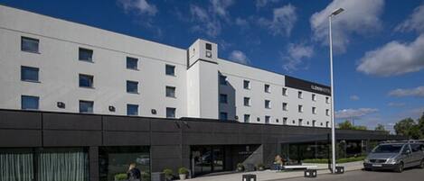Um edifício de hotel moderno com fachada plana, várias janelas e área de estacionamento com um carro e uma van.