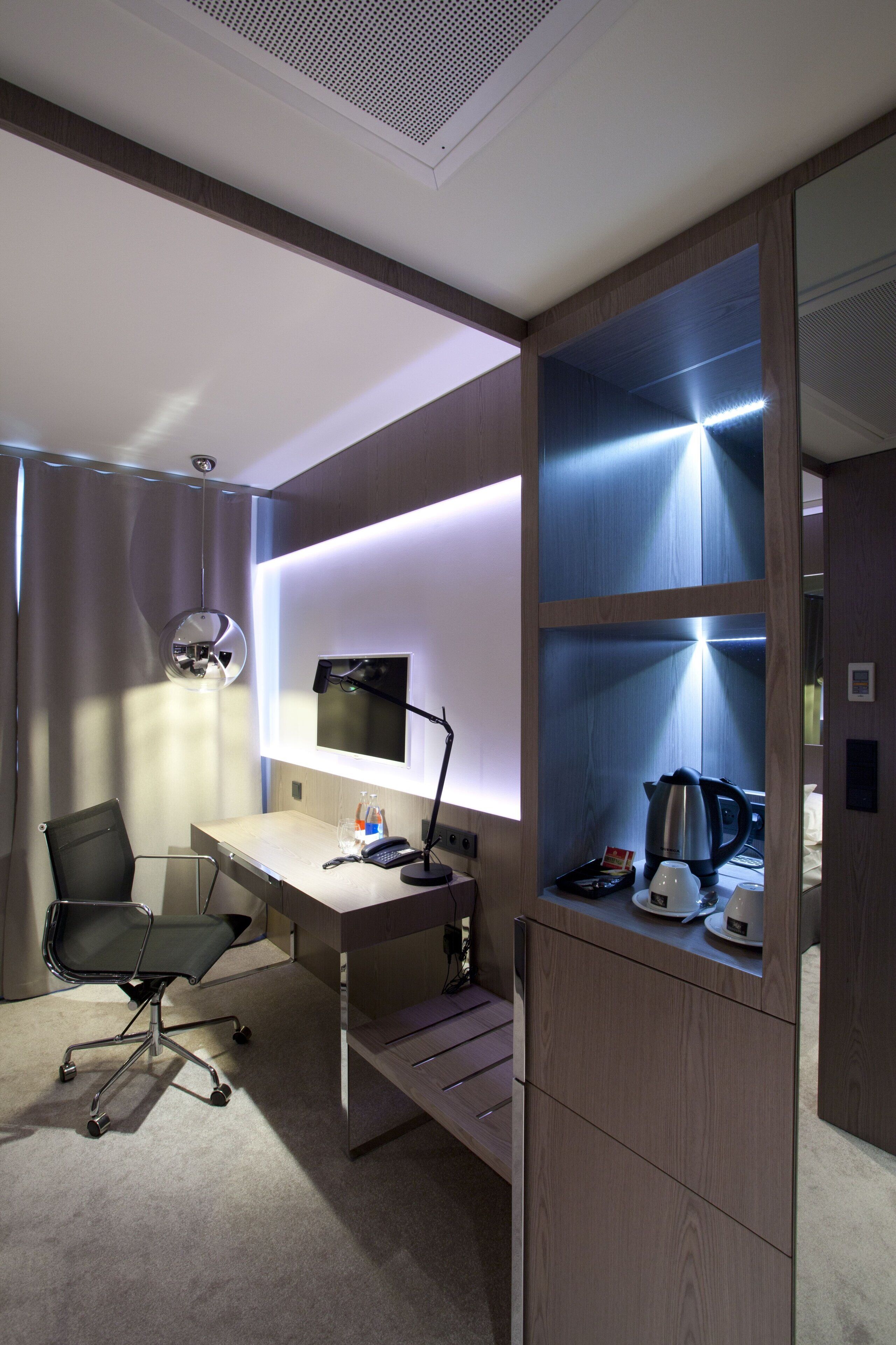 Ein modernes Hotelzimmer mit Schreibtisch, Stuhl und einer integrierten Regaleinrichtung mit Wasserkocher und Kaffeemaschine.