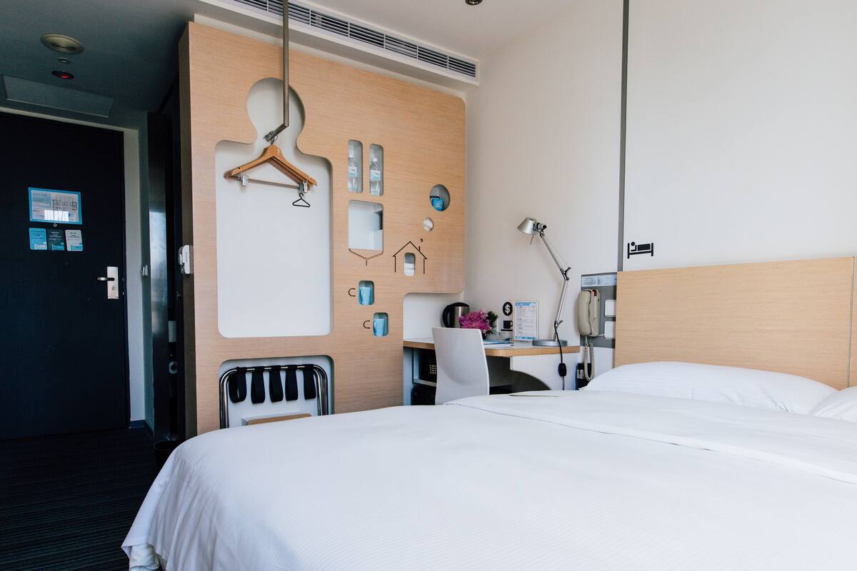A hotel room with a bed, a desk, a chair, a hanging coat rack, and a phone.