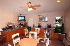 A kitchen with wooden cabinets, a dining table with chairs, a ceiling fan, a television, and a window with plants.