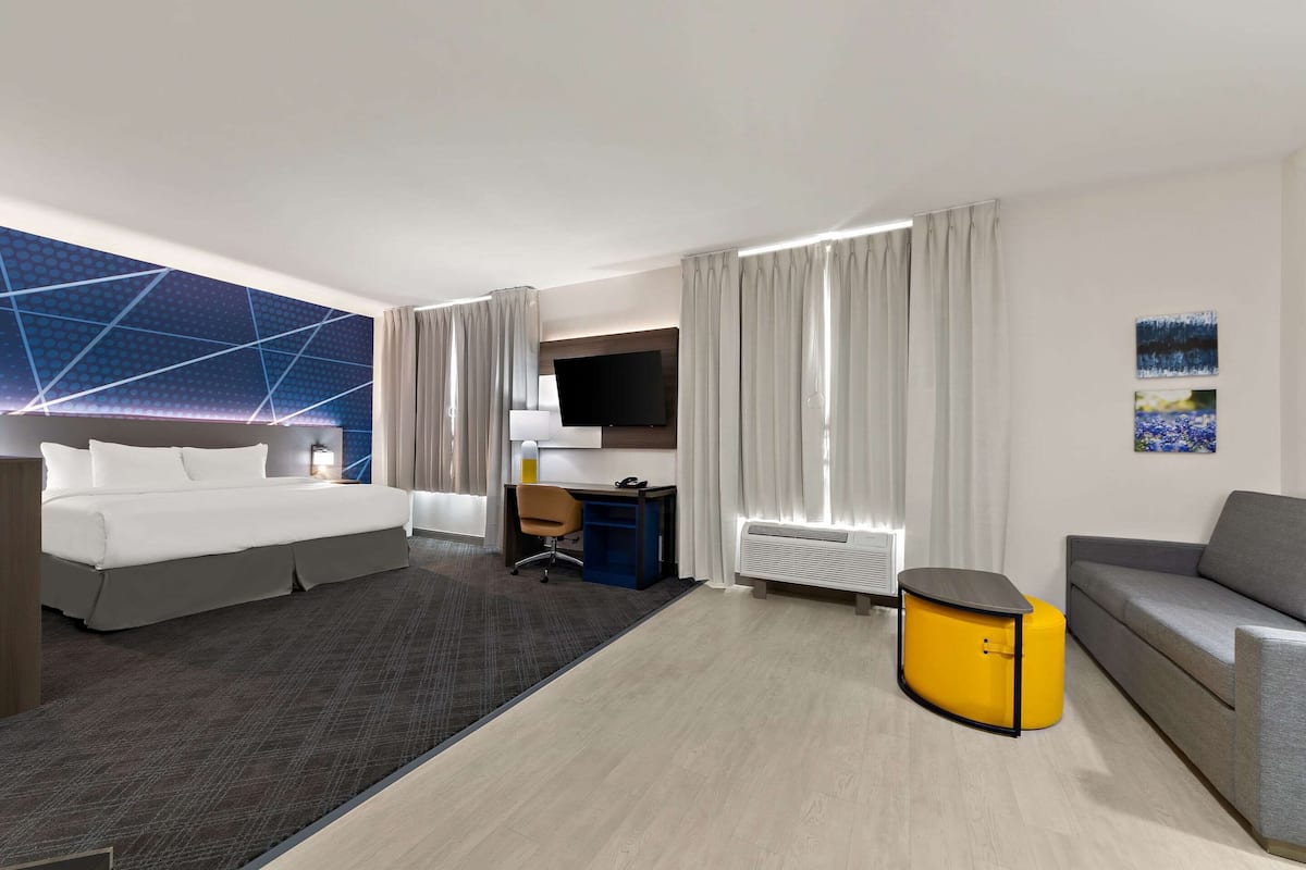 A modern hotel room with a large bed, a desk with a chair, a sofa, a small yellow side table, and a flat-screen TV.