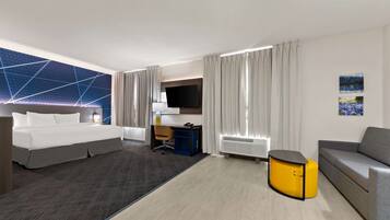 A modern hotel room with a large bed, a desk with a chair, a sofa, a small yellow side table, and a flat-screen TV.