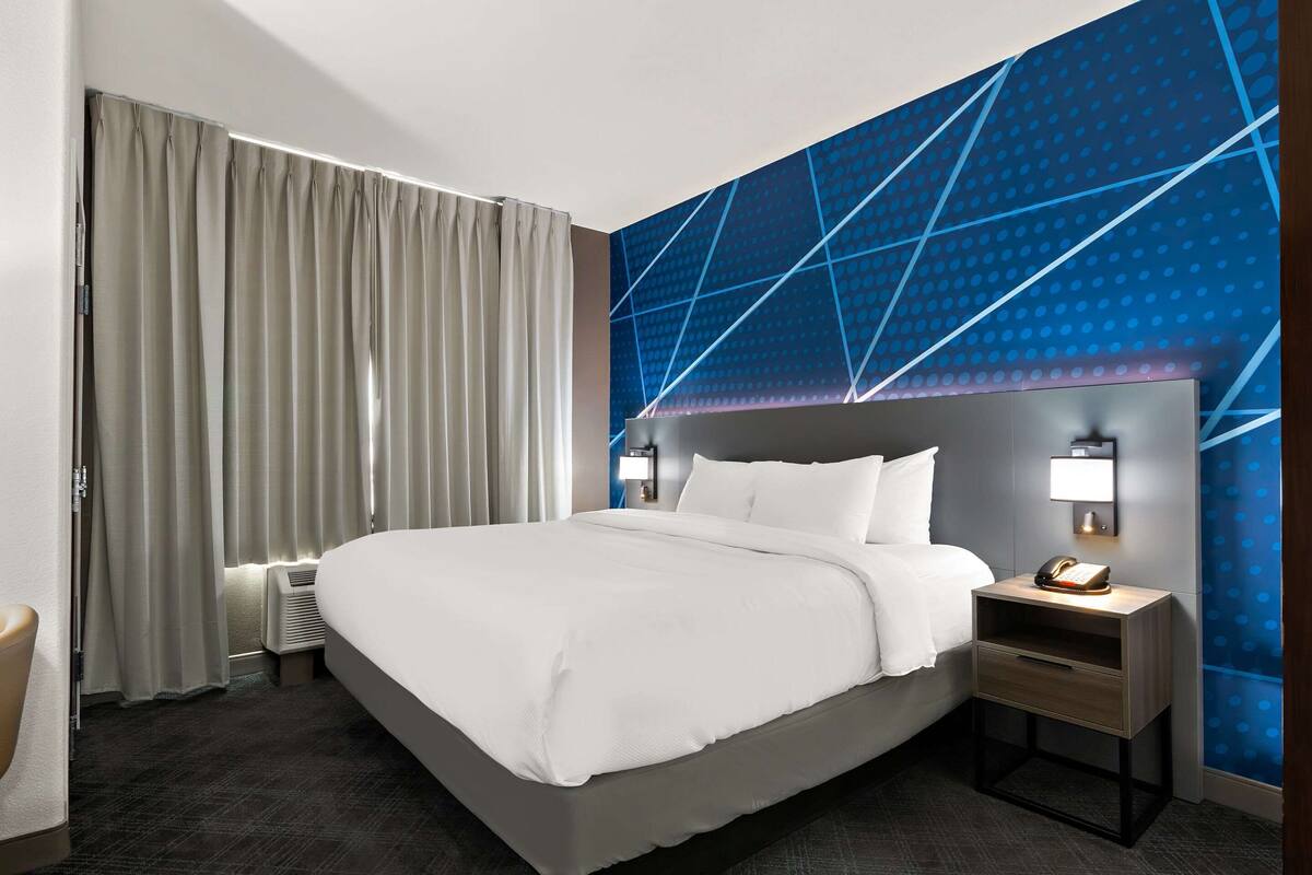 A modern hotel room with a large bed, a nightstand with a lamp, and a geometric-patterned wall.