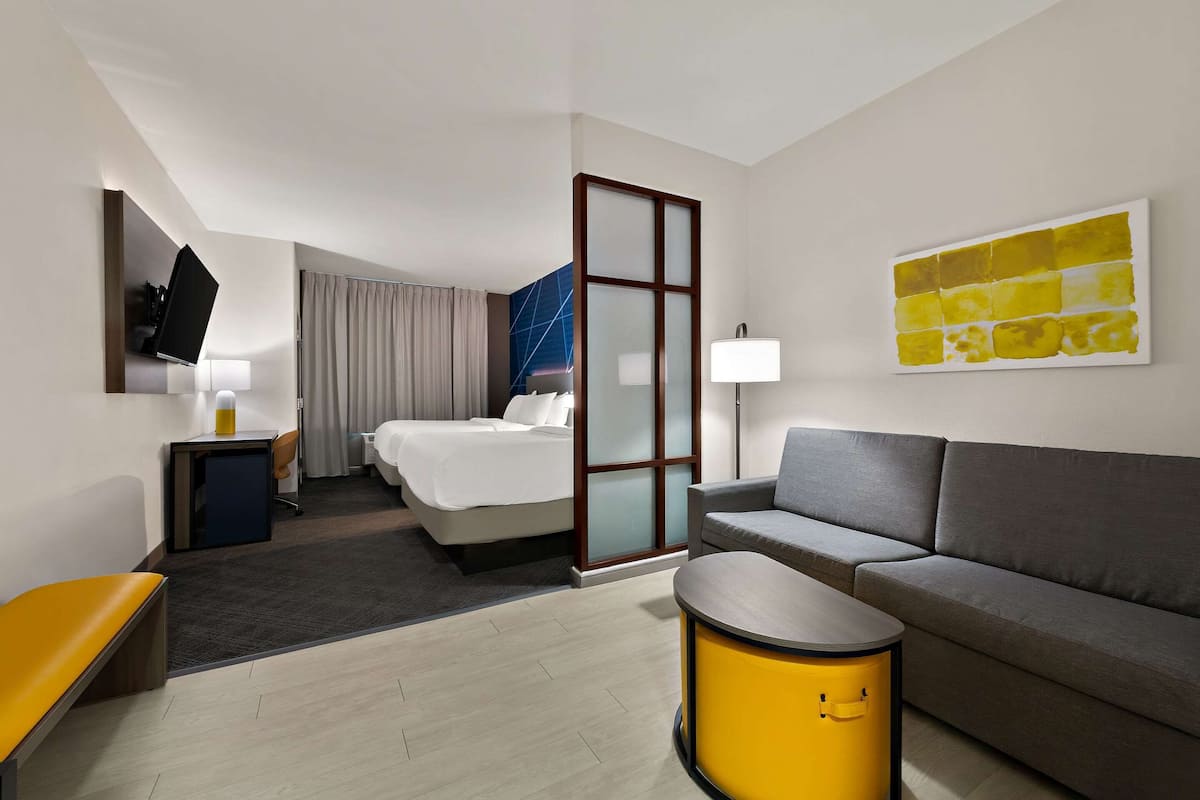 A modern hotel room with a bed, a sofa, a small table, a TV, and a painting on the wall.