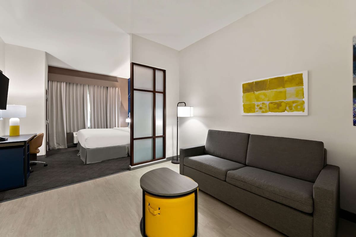 A hotel room with a bed, a sofa, a desk with a lamp, a small yellow stool, and a painting on the wall.