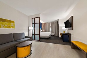 Une chambre d’hôtel comprenant un canapé, une petite table ronde, un lit, un bureau avec une chaise et un téléviseur fixé au mur.