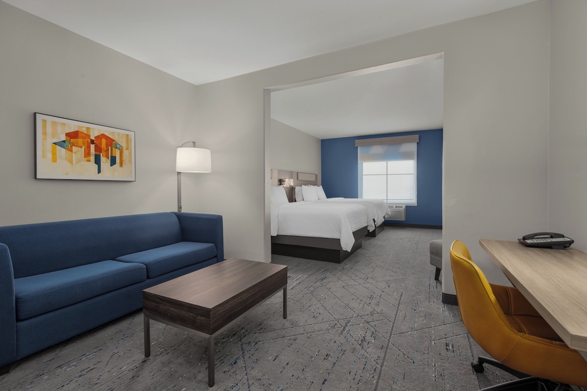 A hotel room with a blue sofa, a wooden table, a bed with white bedding, a desk with a chair, and a painting on the wall.