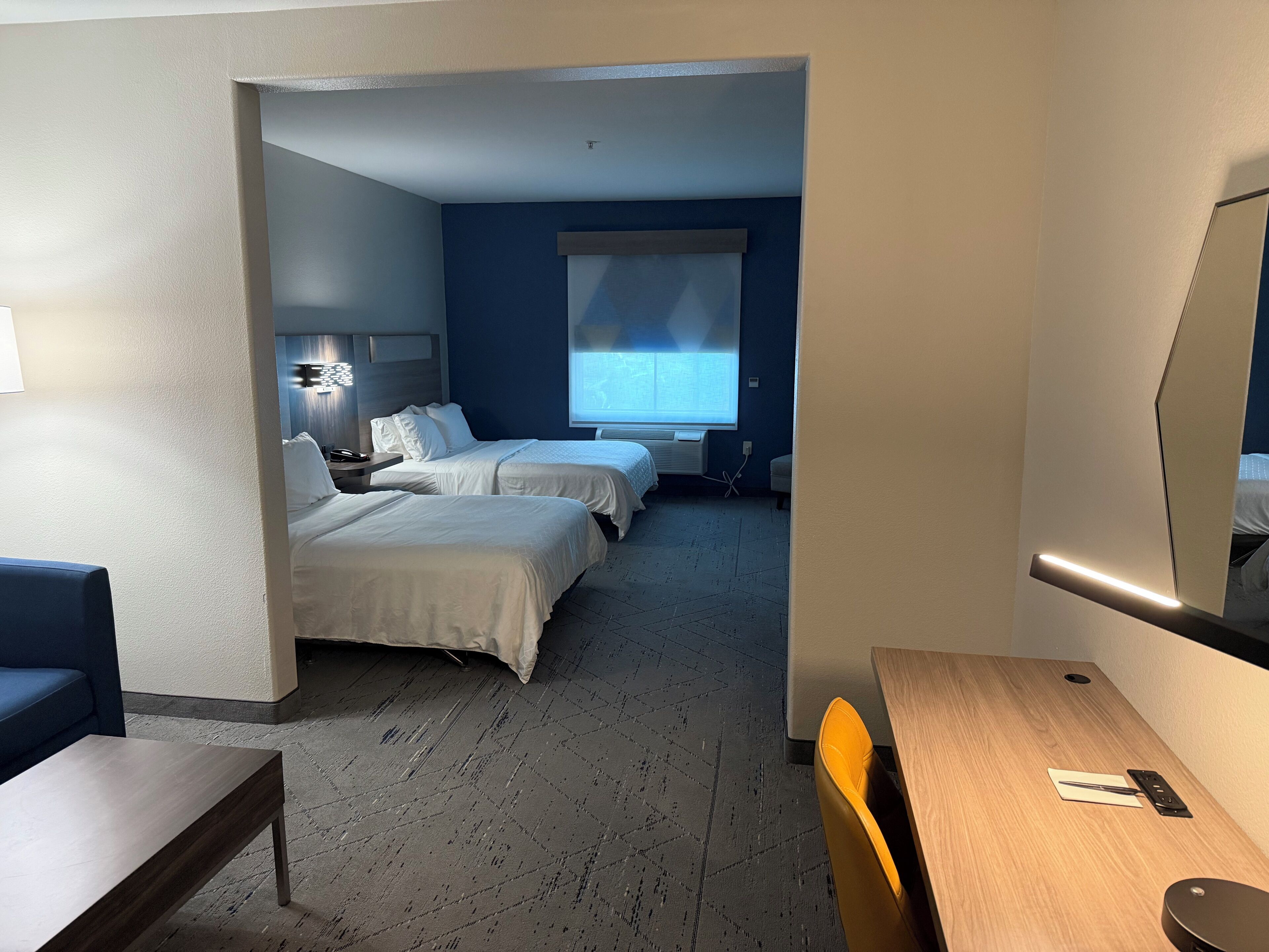 Habitación de hotel con dos camas, un escritorio, una silla, una mesita de noche, una lámpara, una ventana con persianas y un espejo adosado a la pared.