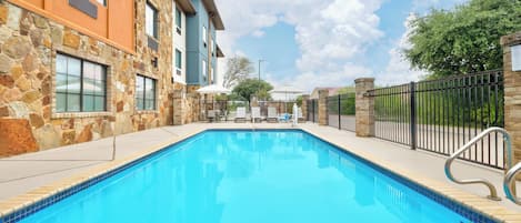 A swimming pool with stone walls, a clear blue sky, and a fenced area with umbrellas.