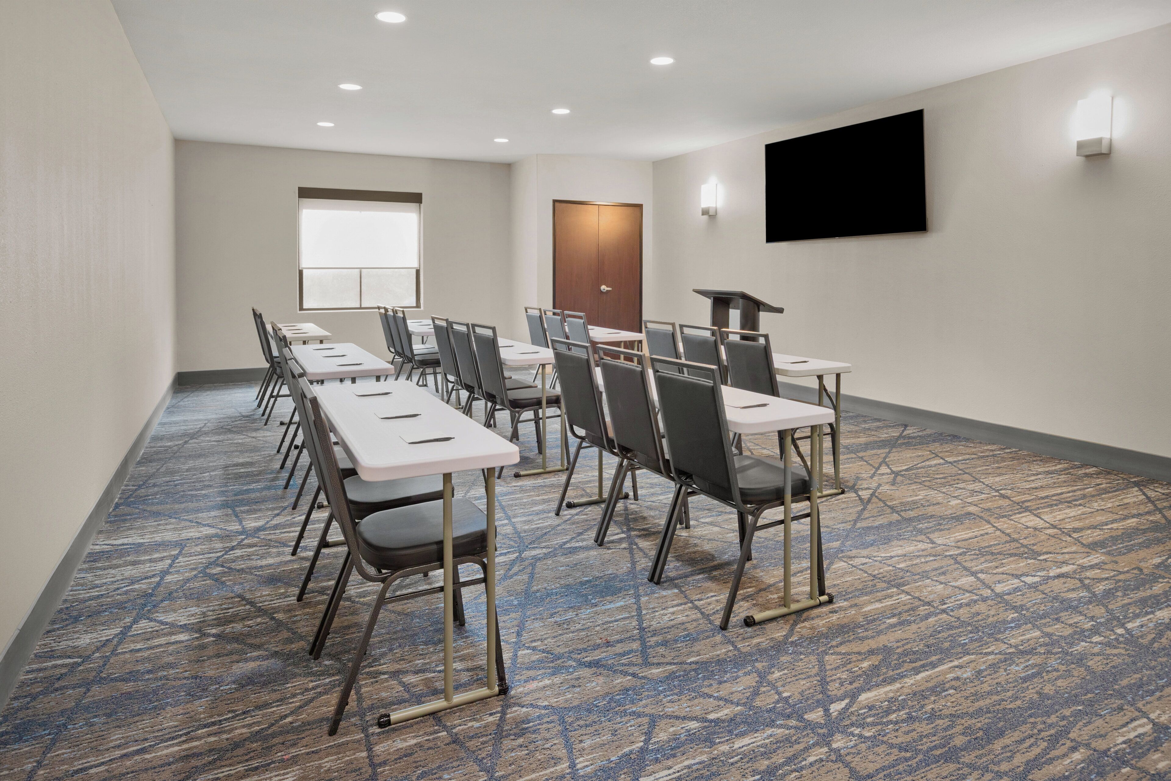 A conference room with tables and chairs, a door, a mounted TV, and a carpeted floor.