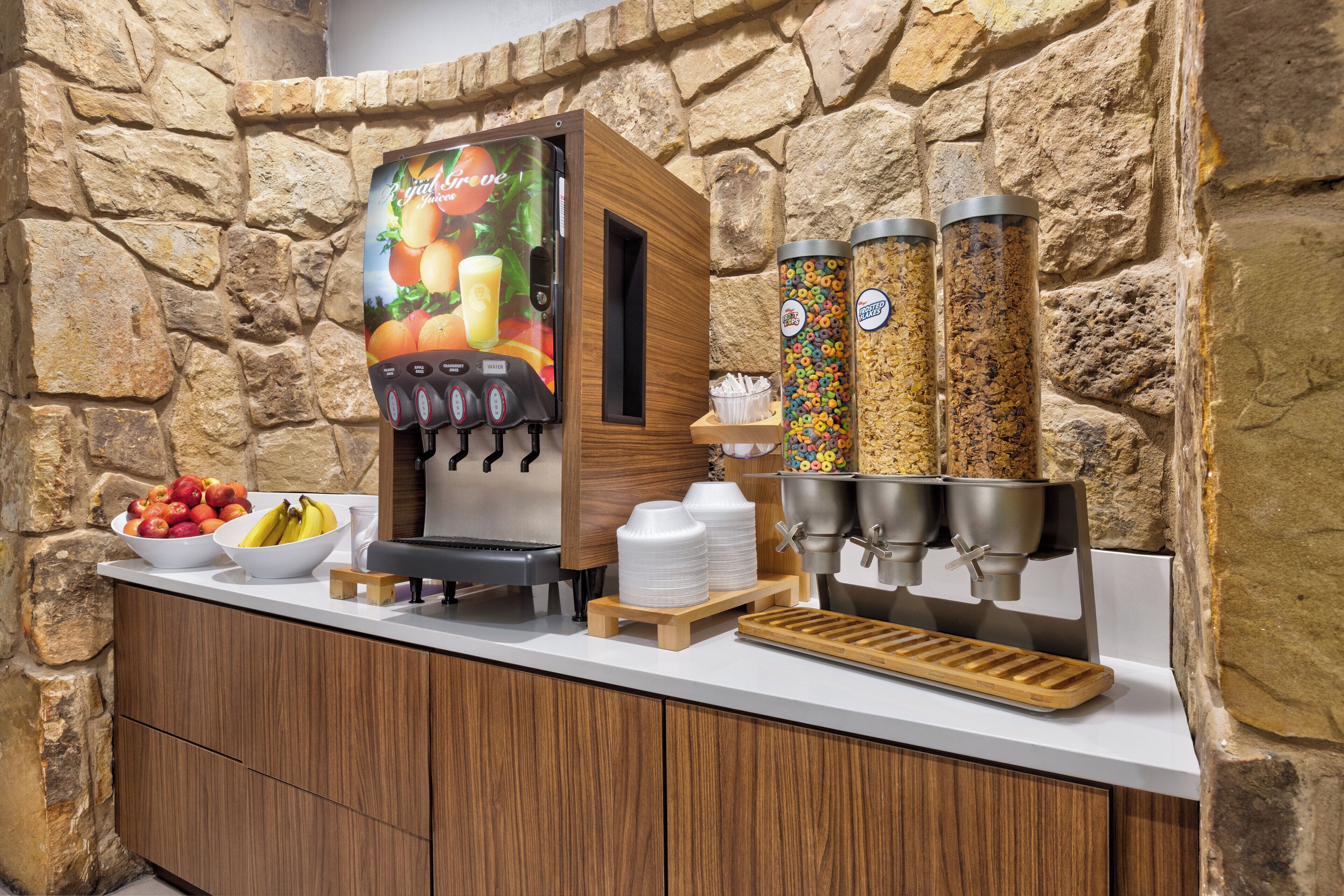 A self-serve beverage station with a fruit juice dispenser, two large condiment dispensers, and a fruit bowl.