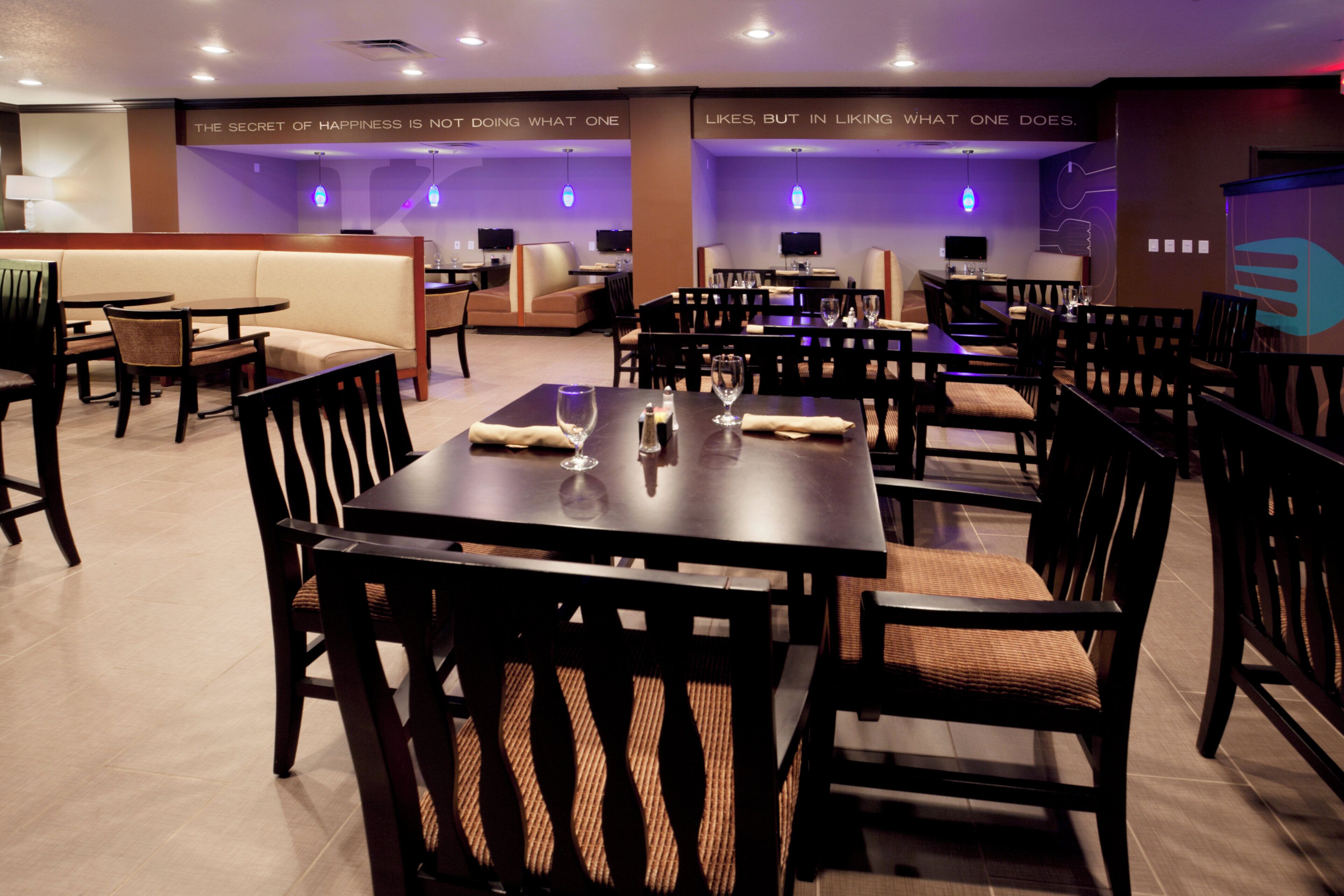 A restaurant with multiple dining tables and chairs, some with cushions. There are wall-mounted televisions and a sign with a motivational quote.