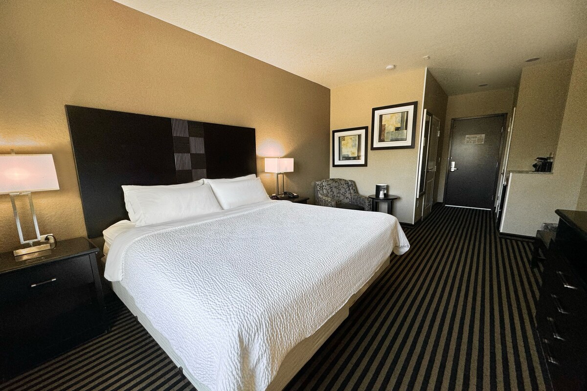 Una habitación de hotel con una cama grande, una mesita de noche con lámpara, una silla, un escritorio y una puerta.