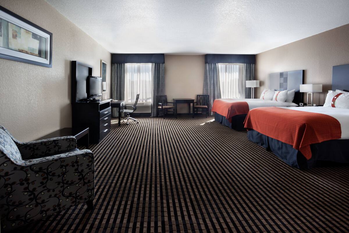Ein Hotelzimmer mit zwei Betten, einem Schreibtisch, einem Sessel, einem Fernseher und einem gemusterten Teppich.