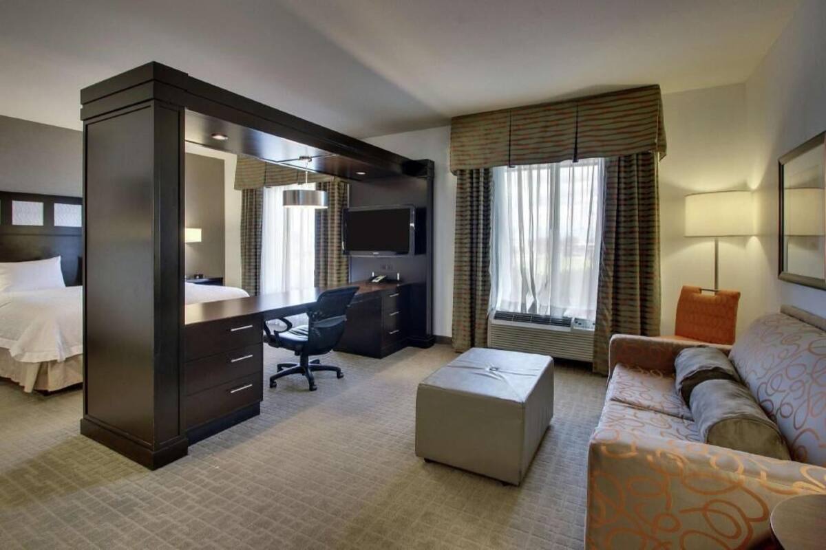 Habitación de hotel con cama, escritorio y silla, sofá, televisor y ventana con cortinas.