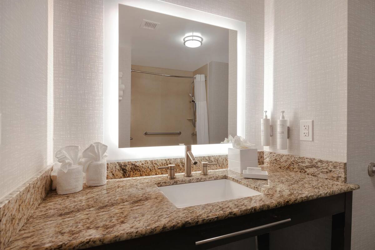 Un baño con encimera de granito, un lavamanos blanco, un espejo grande y una ducha visible al fondo.