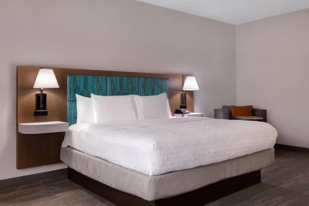 Una habitación de hotel con una cama grande, dos mesitas de noche, una lámpara y un pequeño rincón de descanso con una silla y un cojín.