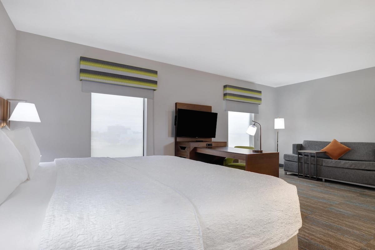 Habitación de hotel con una cama grande, un escritorio con televisor de pantalla plana, un sofá gris y una ventana con persianas.
