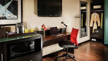 Habitación de hotel con un escritorio de madera, una silla de oficina negra, un televisor de pantalla plana, microondas, cafetera, un jarrón con flores y un armario con bata y zapatos.