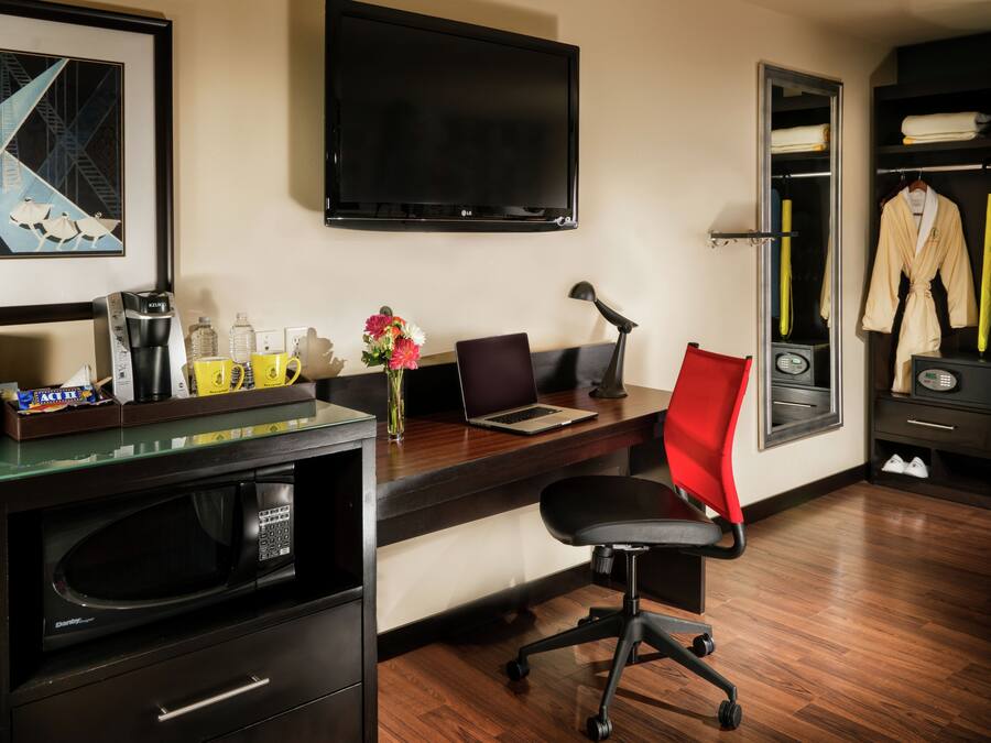 Habitación de hotel con un escritorio de madera, una silla de oficina negra, un televisor de pantalla plana, microondas, cafetera, un jarrón con flores y un armario con bata y zapatos.