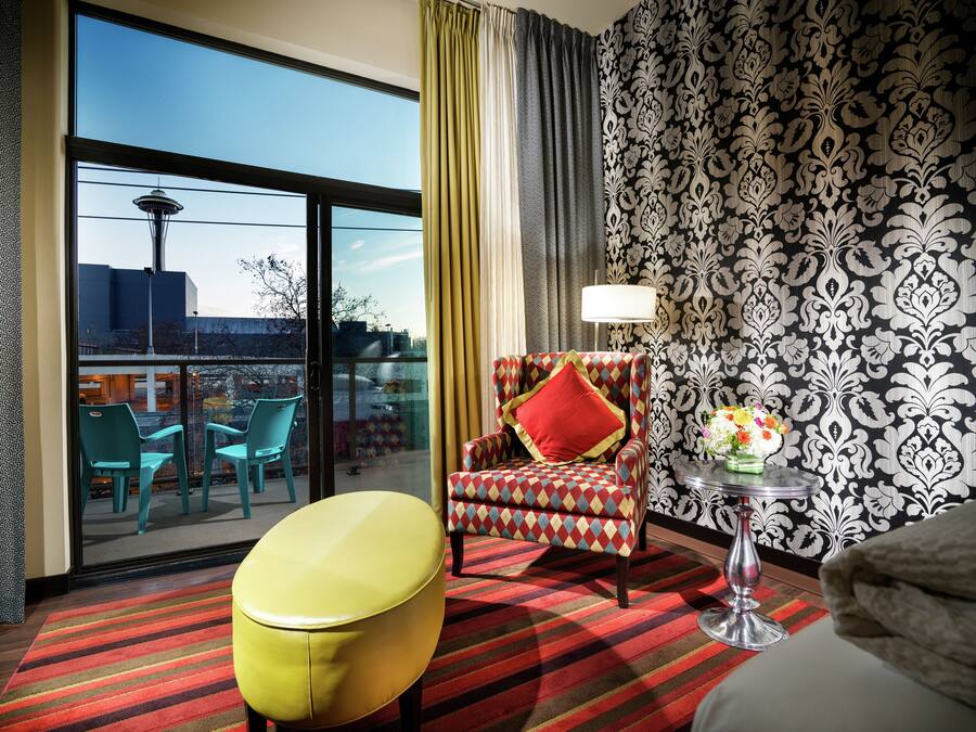 Una habitación de hotel con un ventanal grande, un papel tapiz estampado, un sillón colorido, una mesita con una lámpara y un otoman amarillo.