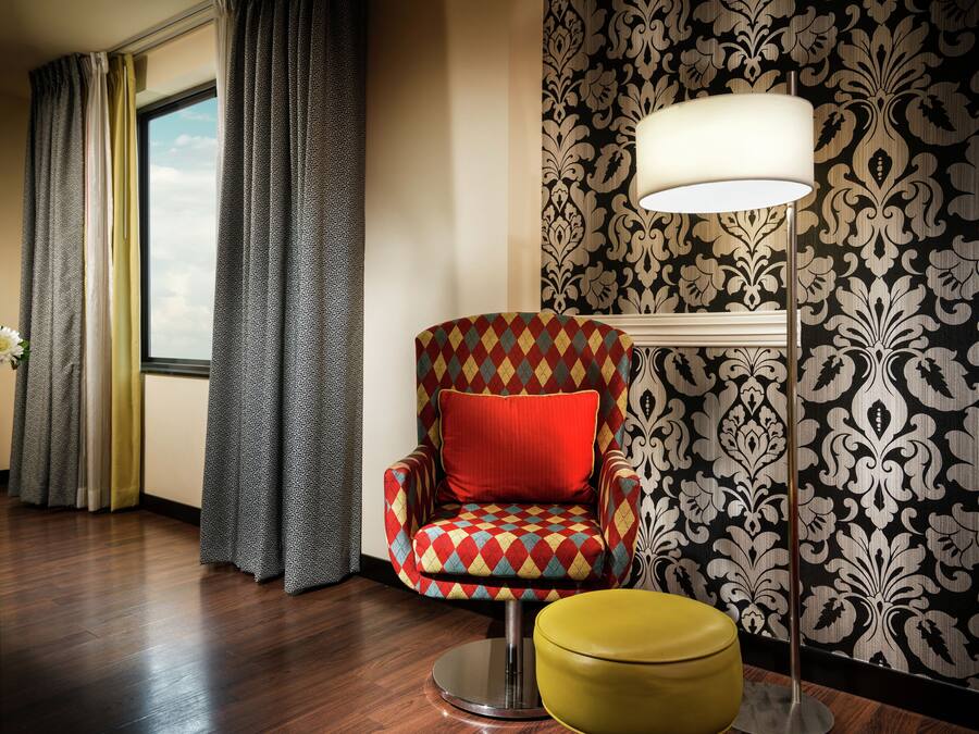 Una habitación con papel tapiz estampado, un sillón rojo y amarillo con cojín, un reposapiés amarillo, una lámpara de pie y una ventana con cortinas.