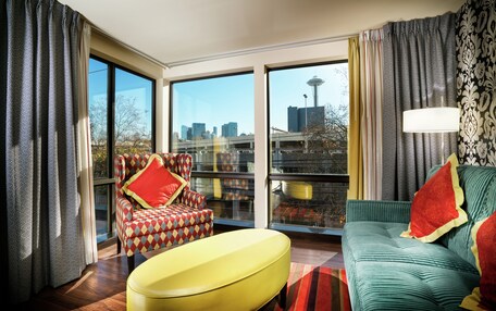 Una sala de estar moderna con ventanas amplias, un reposapiés amarillo, un sofá verde y un sillón estampado.