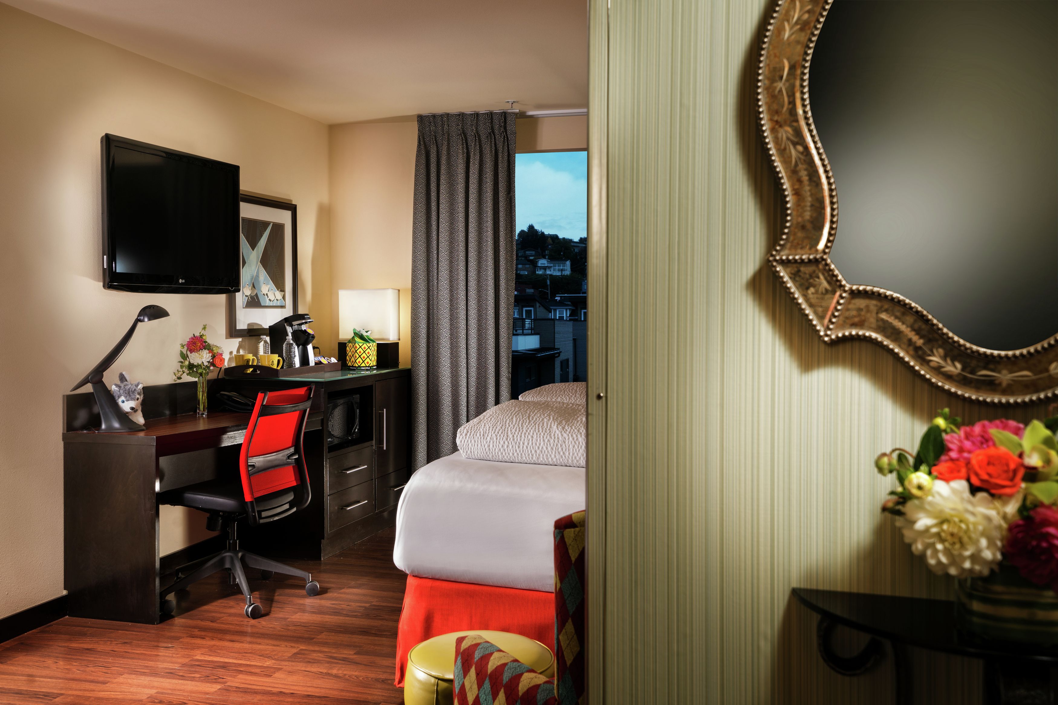 A hotel room with a bed, desk, chair, TV, and a view of the city.