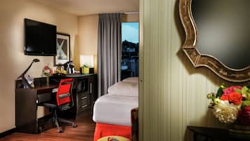 A hotel room with a bed, desk, chair, TV, and a view of the city.