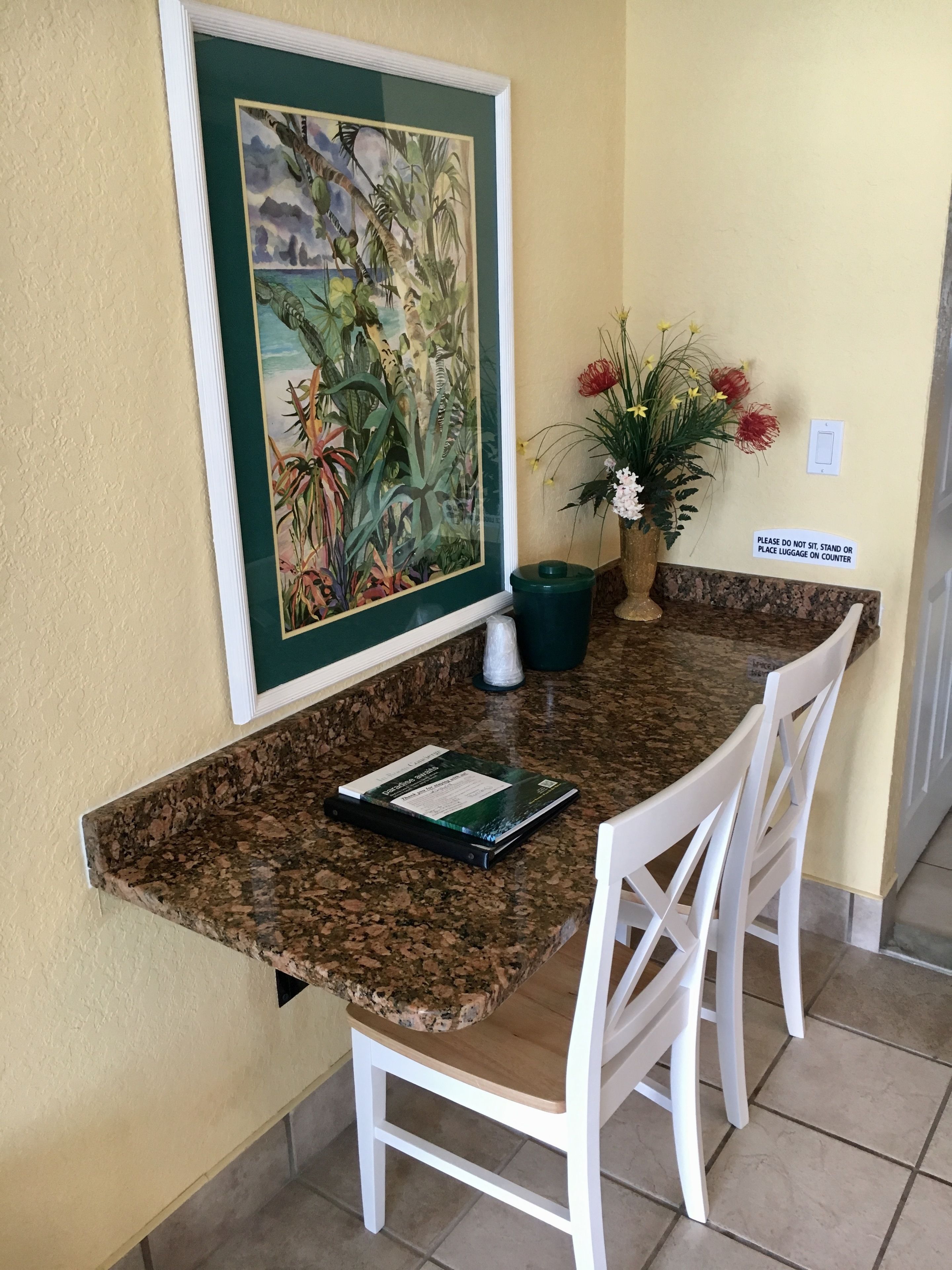 Un coin cuisine doté d’un plan de travail en granit, de chaises blanches, d’un tableau tropical encadré, d’une plante en pot et d’un panneau indiquant qu’il est interdit de s’asseoir ou de poser des bagages sur le plan de travail.