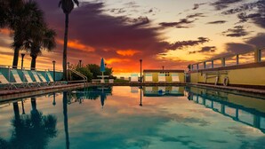 Una piscina con sillas de descanso y palmeras, reflejando el atardecer.