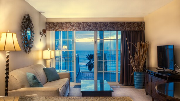 Una sala de estar con sofá, mesa de centro y televisor, una ventana grande con vista al océano, y un espejo decorativo en la pared.