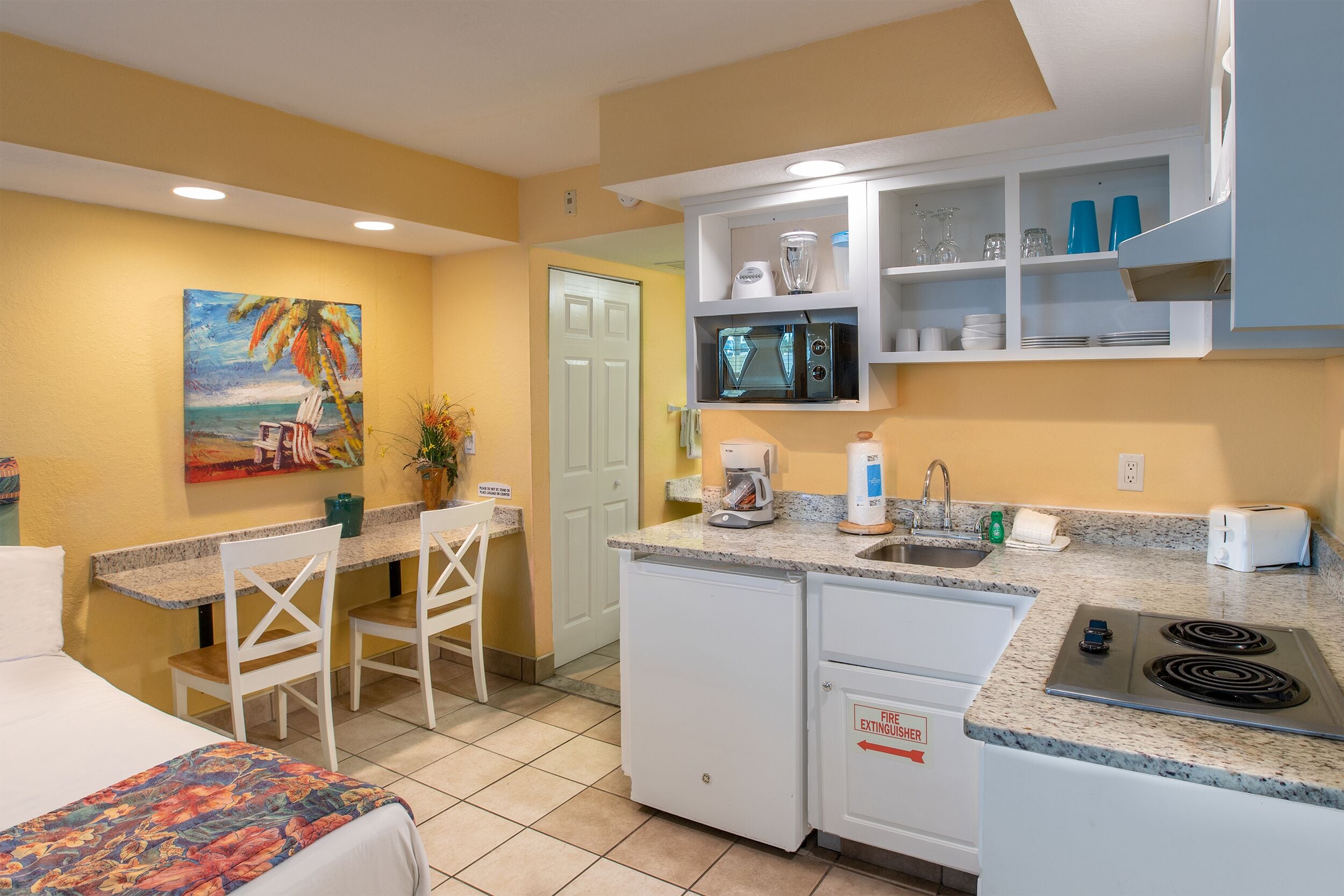 A compact kitchen with a microwave, toaster, and sink. There is a dining area with chairs and a table. A painting of a beach scene is on the wall.