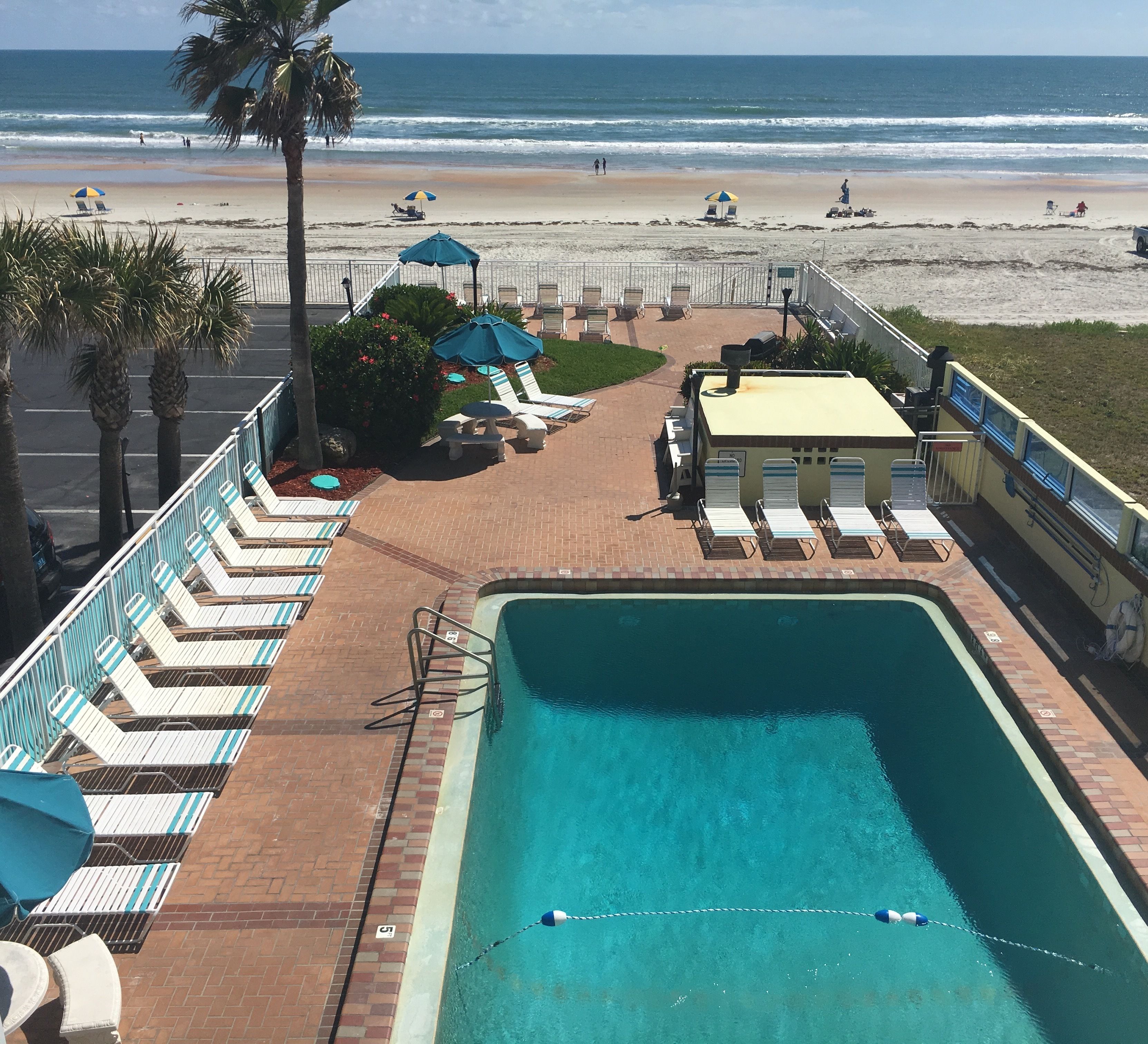 Uma área de piscina com espreguiçadeiras e guarda-sóis, uma praia ao fundo e vista para o oceano.