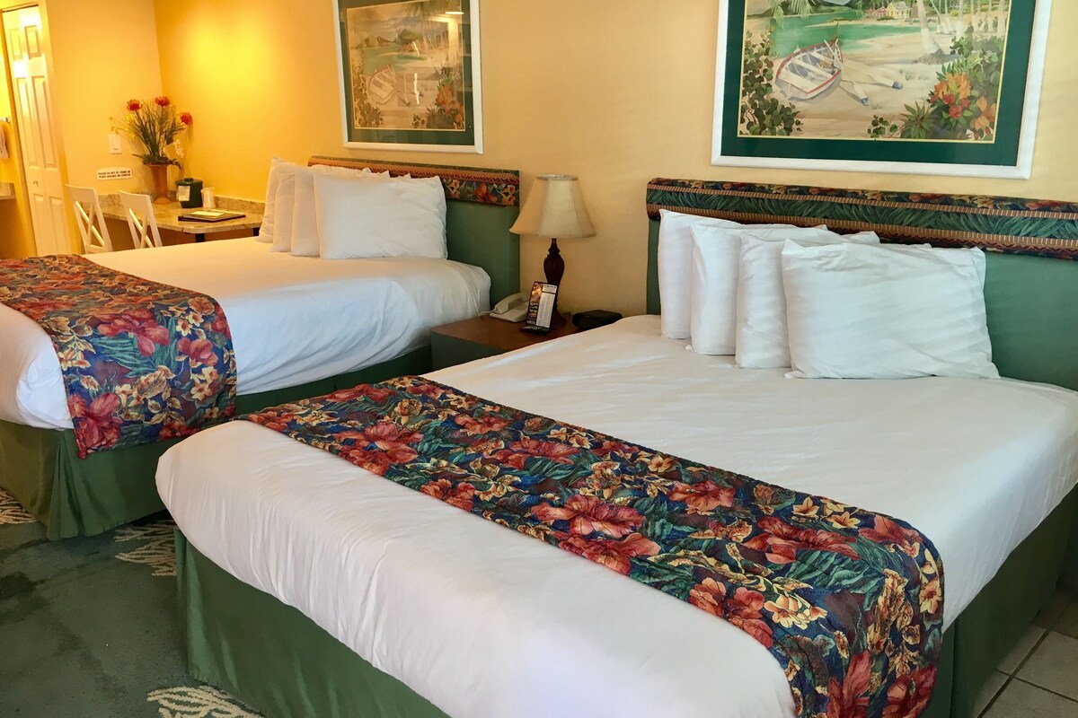 A hotel room with two beds, a desk, a chair, a lamp, and a painting on the wall.