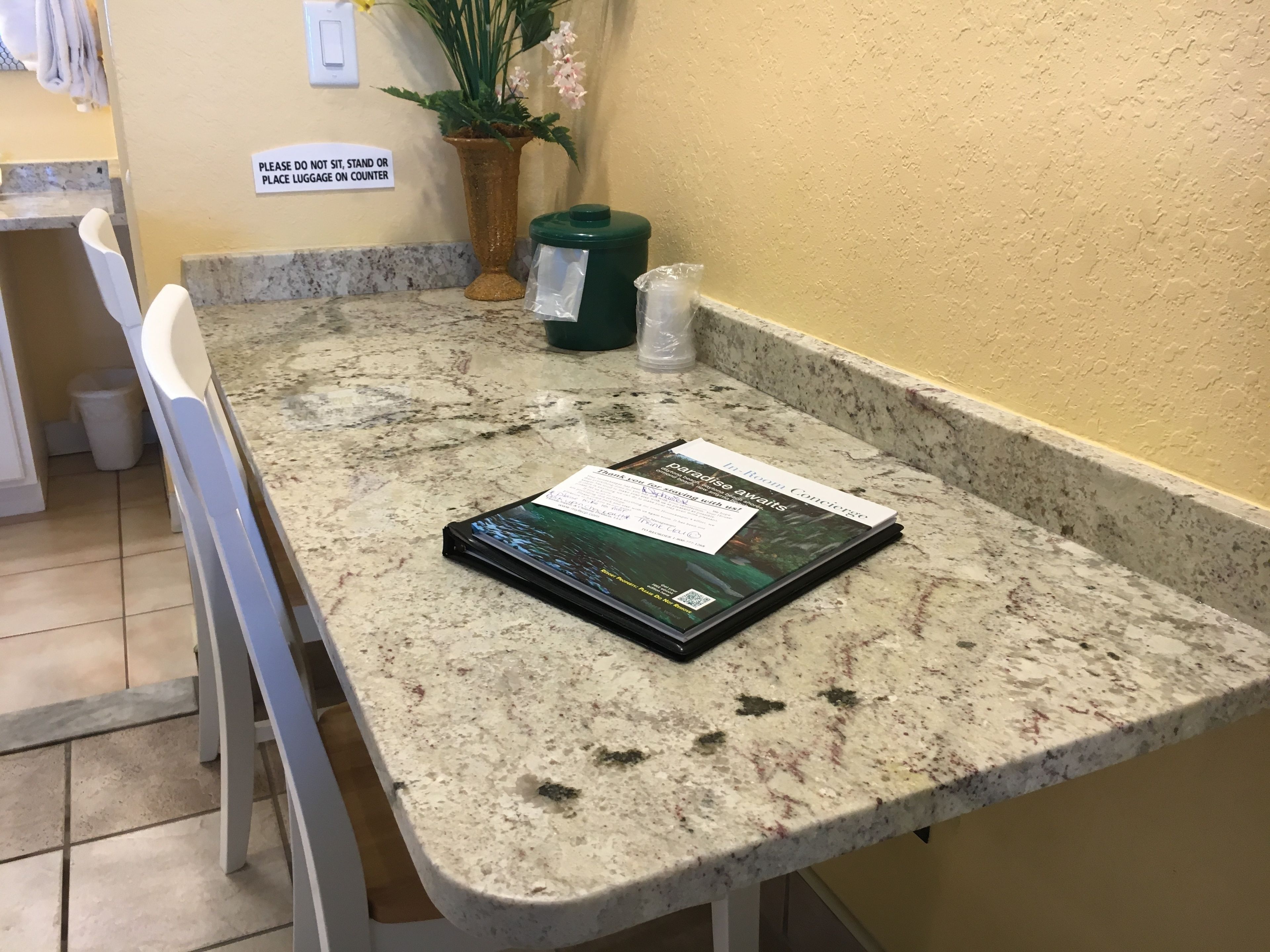 A kitchen counter with a book titled "Paradise Awaits" and a sign instructing not to sit, stand, or place luggage on the counter.