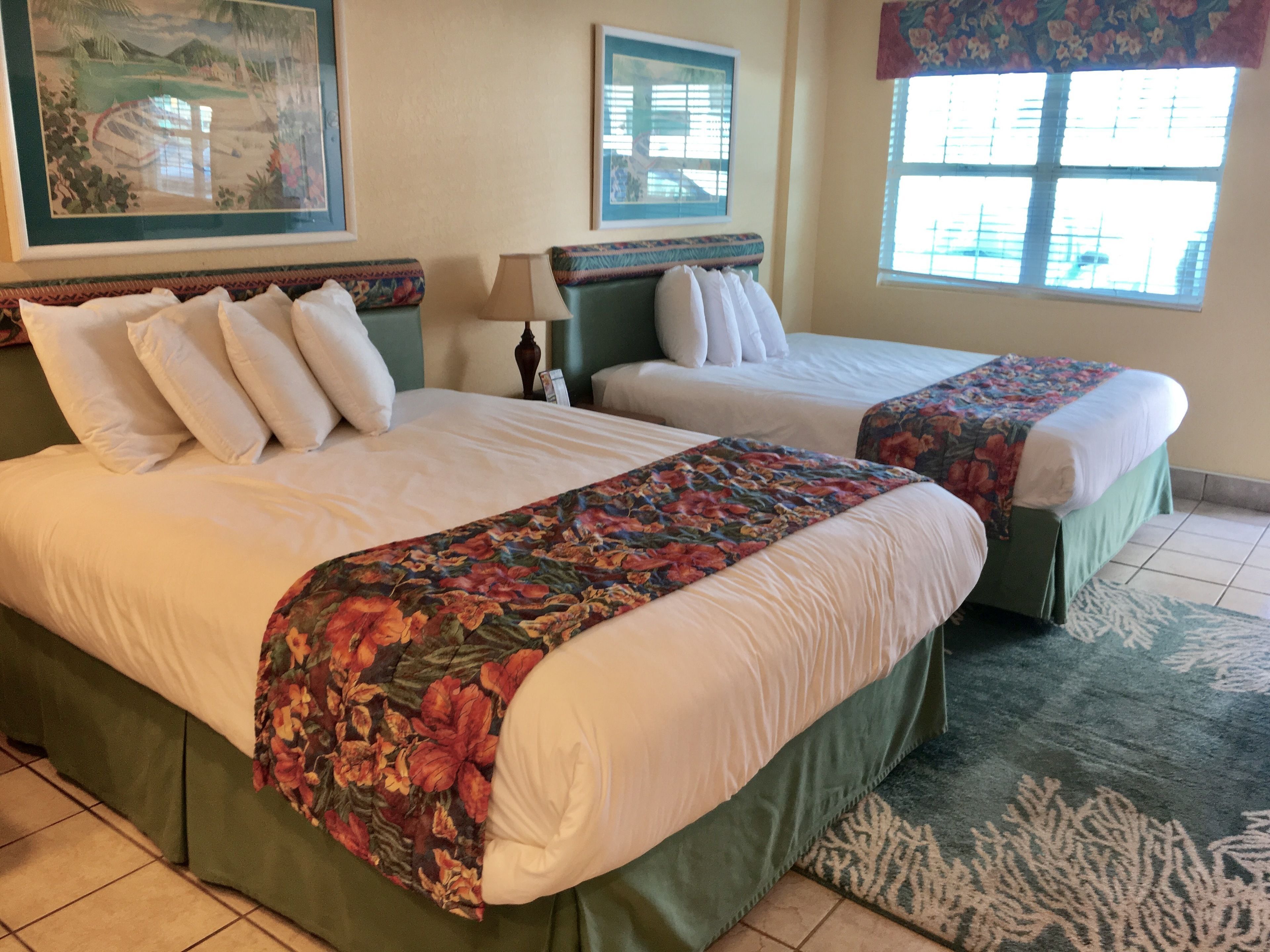 A hotel room with two beds, a green bed skirt, a floral bedspread, a green headboard, a window with blinds, and a framed picture on the wall.