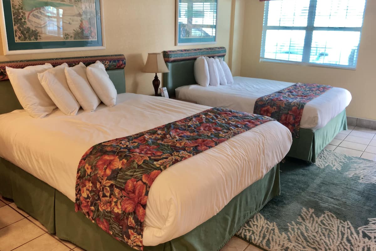 A hotel room with two beds, a green bed skirt, a floral bedspread, a green headboard, a window with blinds, and a framed picture on the wall.