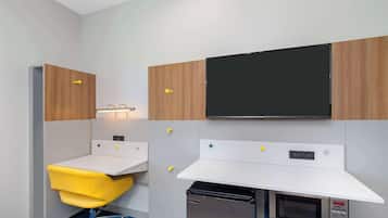 A compact hotel room with a kitchenette, a small desk, a yellow chair, and a flat-screen TV mounted on the wall.