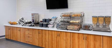 A buffet counter with various food dispensers, a television mounted on the wall, and a tiled floor.