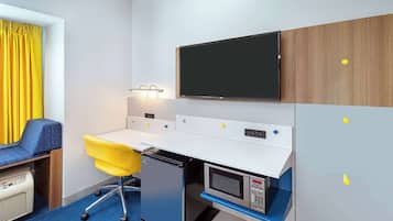Habitación de hotel compacta con kitchenette, televisión de pantalla plana, pequeño escritorio con silla y ventana con cortinas amarillas.