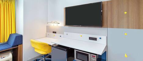 A compact hotel room with a kitchenette, a flat-screen TV, a small desk with a chair, and a window with yellow curtains.